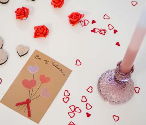 a white table with plastic hearts scattered on top. A pink candle is to the right and a homemade Valentine's Day card is on the left