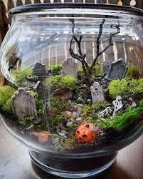 A large, glass fishbowl is shown filled with dirt, headstones, pumpkins, and moss.